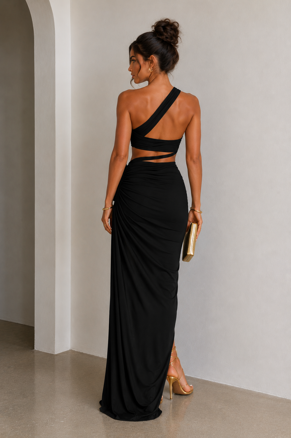 Woman in a black one-shoulder dress standing against a plain wall.