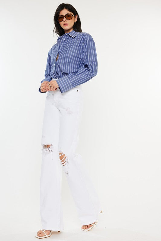 Model is wearing White High Rise Flare Jeans with white heels and a blue stripped shirt, the model is also wearing chic sunglasses and silver rings. Side front view. 