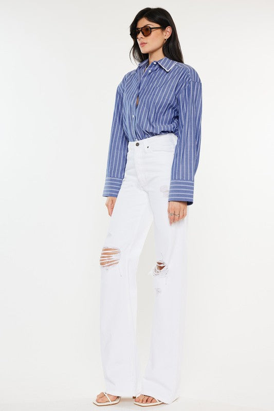 Model is wearing White High Rise Flare Jeans with white heels and a blue stripped shirt, the model is also wearing chic sunglasses and jewelry. Side front view.