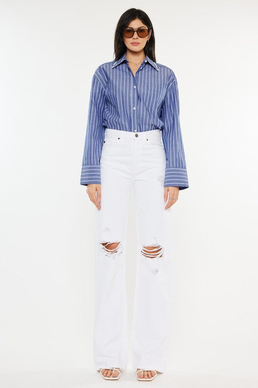 Model is wearing White High Rise Flare jeans with a blue striped shirt, sunglasses and white sandals. Front view. 