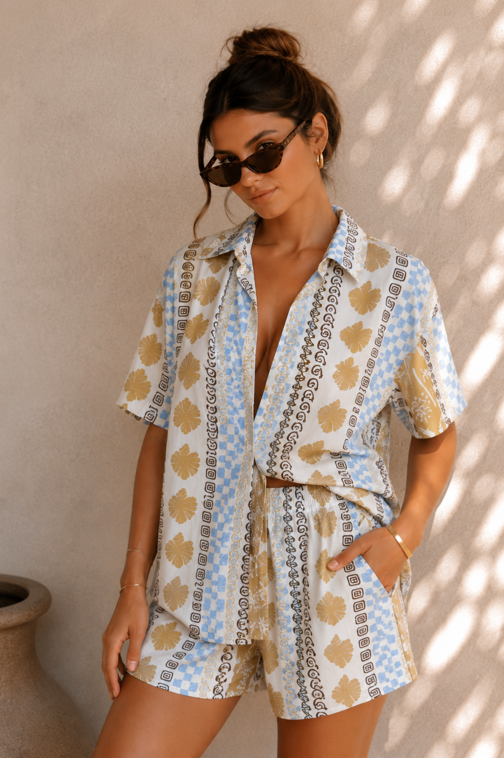 Woman wearing a patterned shirt and shorts set against a neutral wall.
