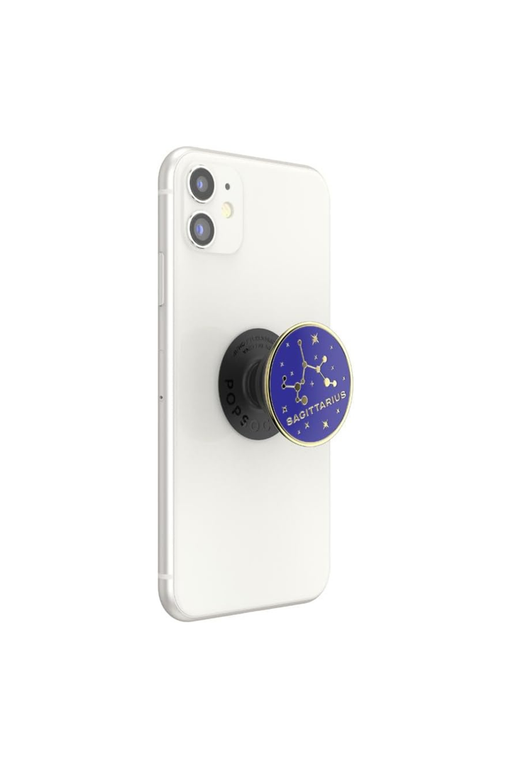 A white smartphone shown from the back with a black circular pop socket attached near the lower center. The pop socket has a blue face with gold constellation line art and the words &
