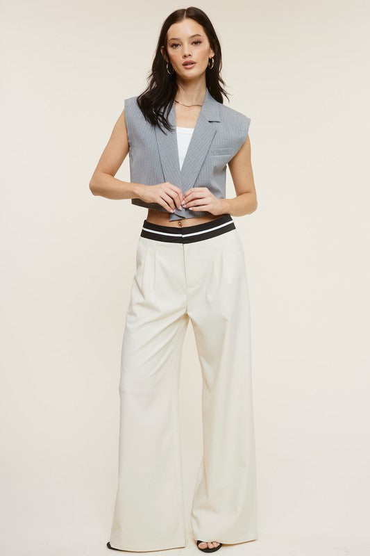 Model is wearing Cream Talbot Trousers with a grey vest  with a white top under it and gold hoop earrings 