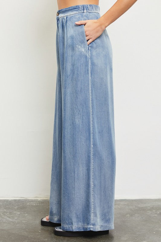 Model is wearing Denim Wide Leg Pants while putting hands in side pockets and wearing black sandals. Side view 