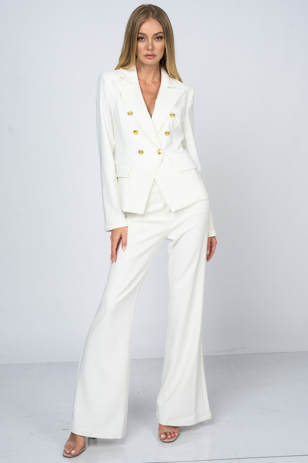A woman modeling a white double-breasted blazer with gold buttons and matching white wide-leg trousers, styled with clear high-heeled sandals against a light gray studio background.