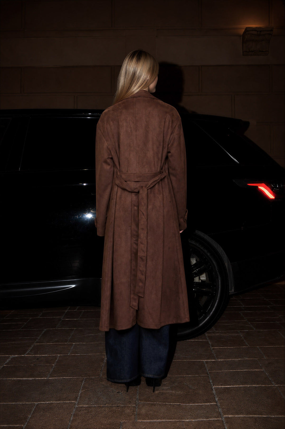 Back view of the dark brown faux suede trench coat with tie belt, styled with dark jeans at night
