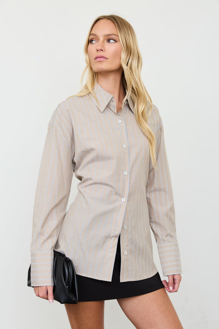 A woman modeling a beige long-sleeve button-up shirt with thin vertical light blue stripes, paired with a black mini skirt and holding a small black clutch bag, set against a plain light gray studio background.
