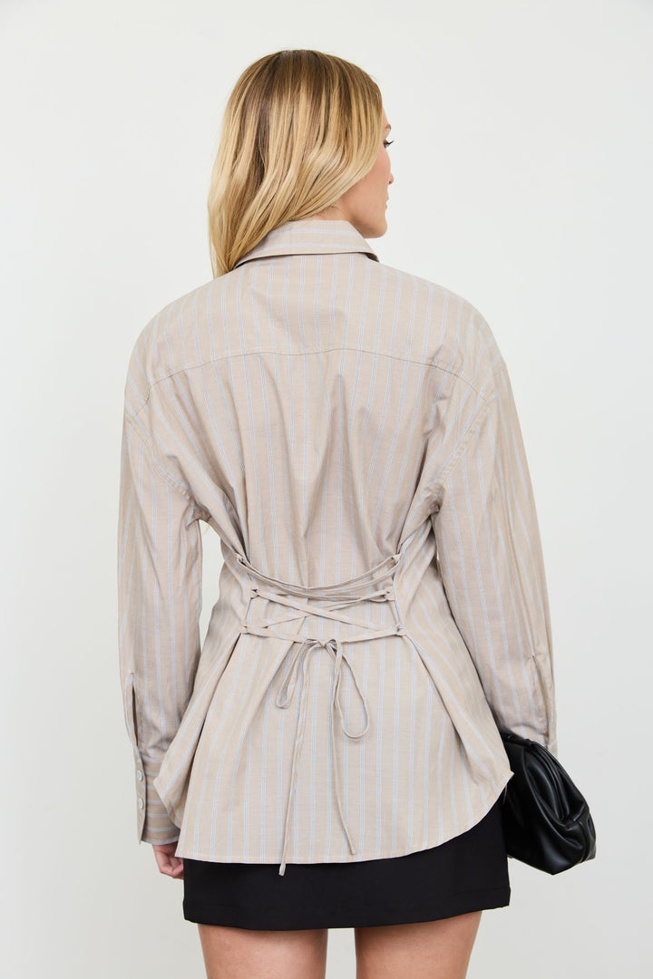 A woman shown from the back wearing a beige and white vertically striped long-sleeve wrap shirt with a tie waist, paired with a black skirt and holding a black clutch bag, against a plain white studio background.