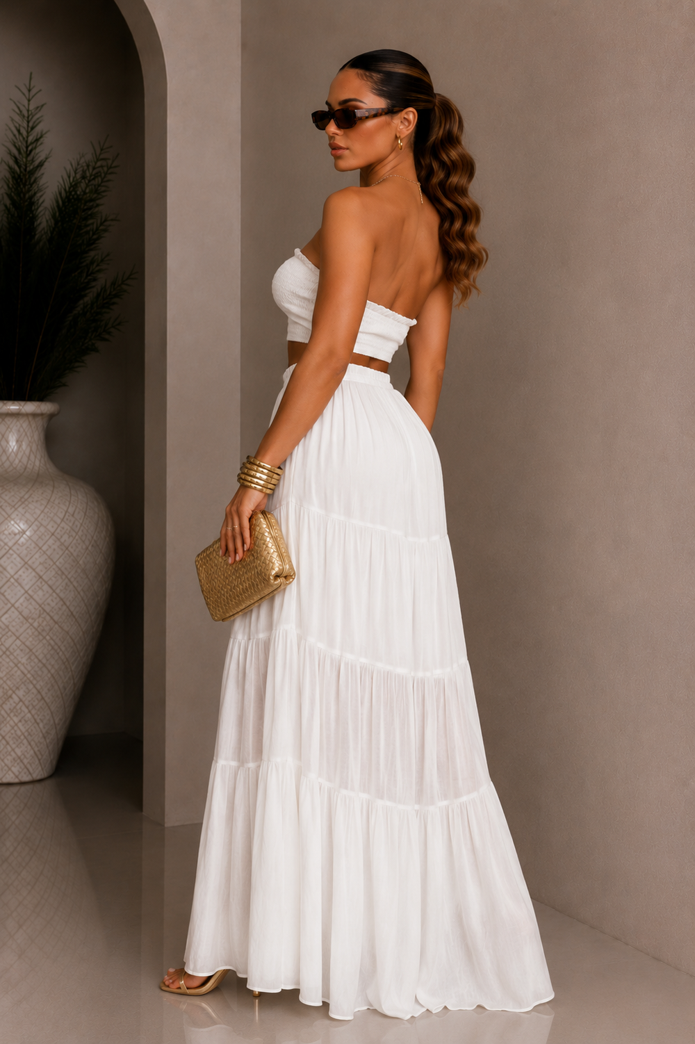 Woman in a white dress holding a gold clutch against a neutral background. White Strapless Top & Maxi Skirt Set. 