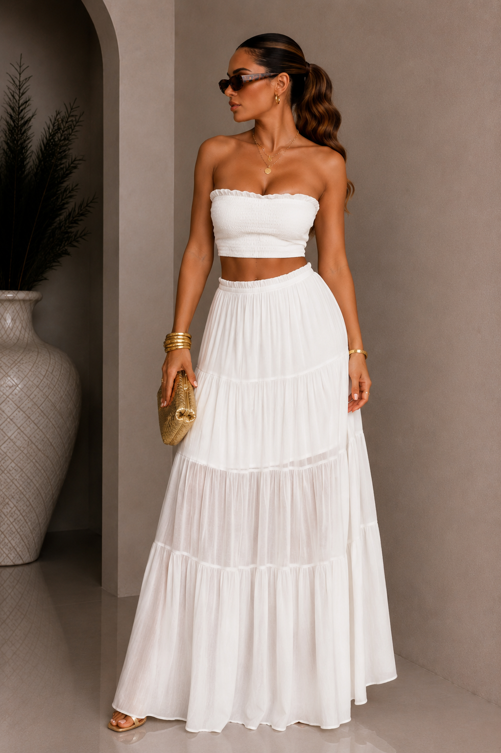 Woman wearing a white strapless top and tiered skirt with gold clutch against a neutral background. White Strapless Top &amp; Maxi Skirt Set. 