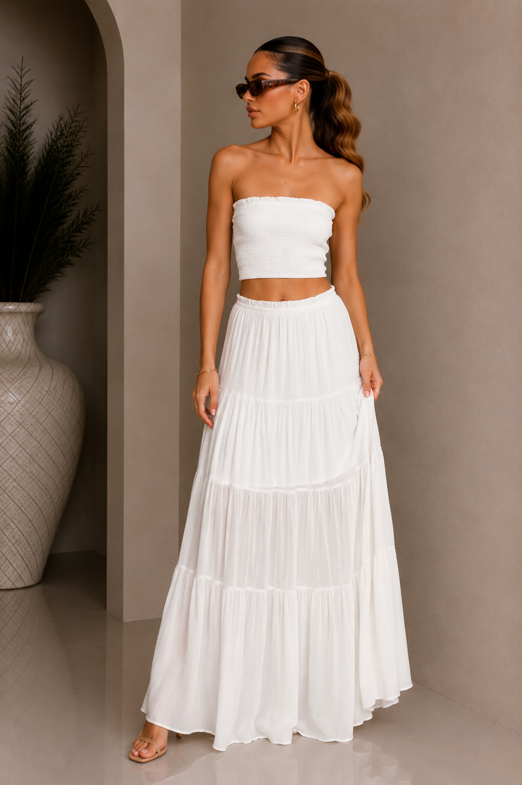 Woman wearing a white strapless top and long skirt standing against a beige wall. White Strapless Top &amp; Maxi Skirt Set.