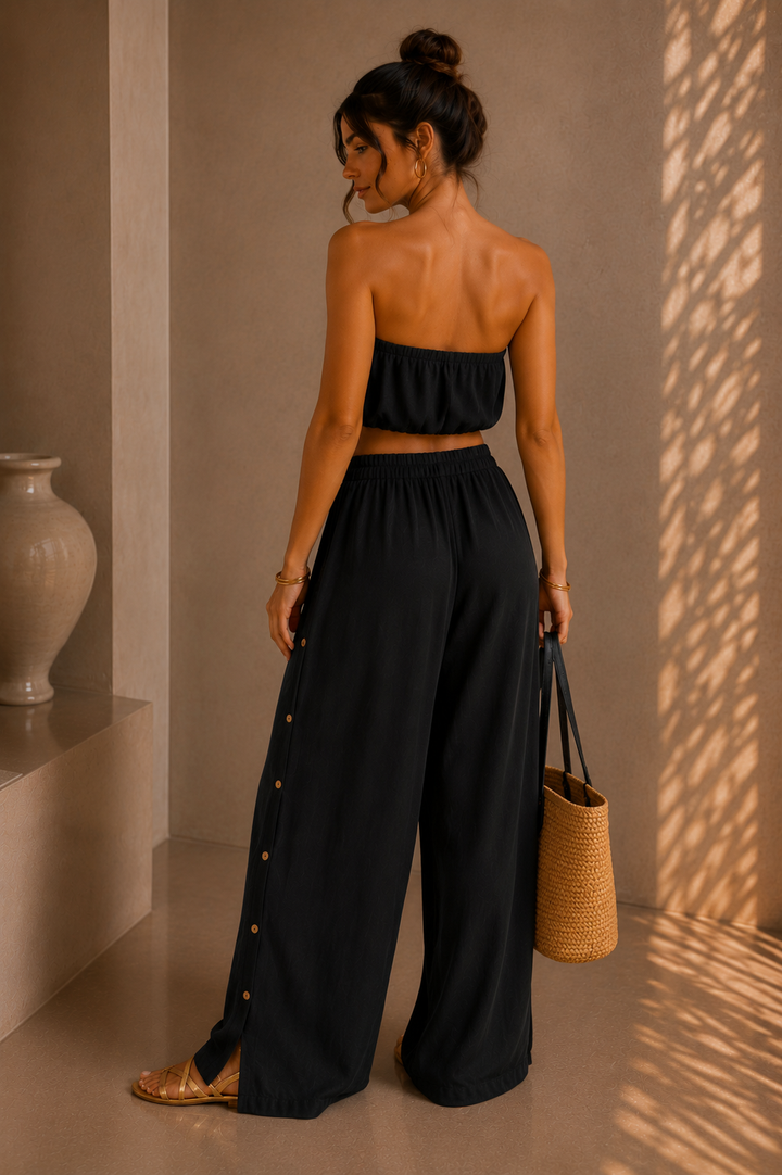 Woman in black outfit holding a woven bag in a sunlit room. Black Strapless Top &amp; Pants Set.