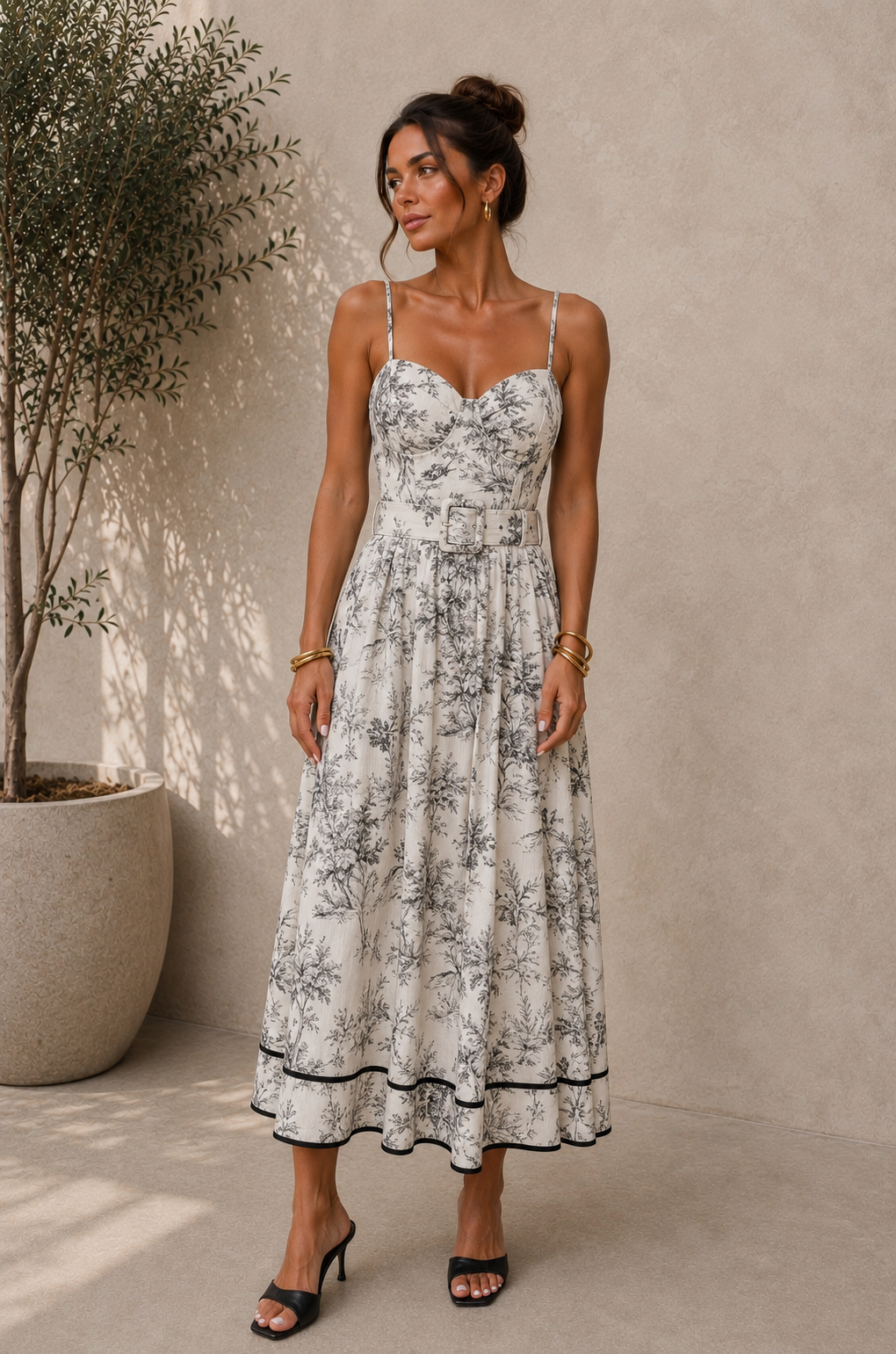 A woman models a sleeveless midi dress with thin straps, a sweetheart neckline, and a black floral toile pattern on a white background. The dress features a matching belt at the waist and a black piped hem, shown against a textured beige wall next to a potted plant.