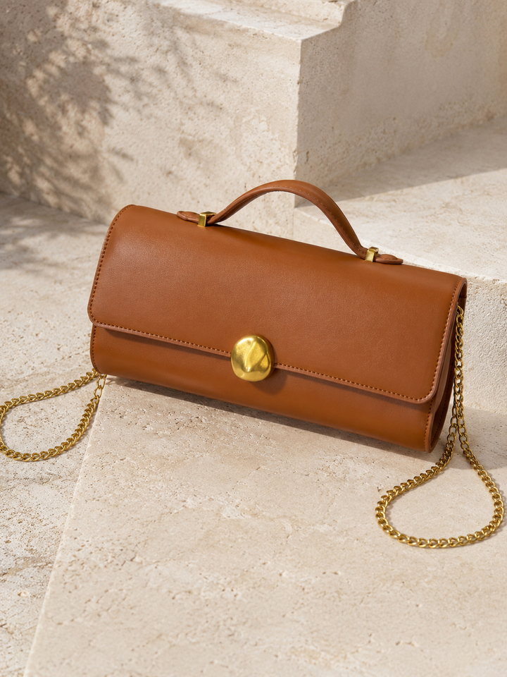 Brown leather handbag with gold chain and clasp on a light stone surface. Tan Top Handle Clutch Bag. 
