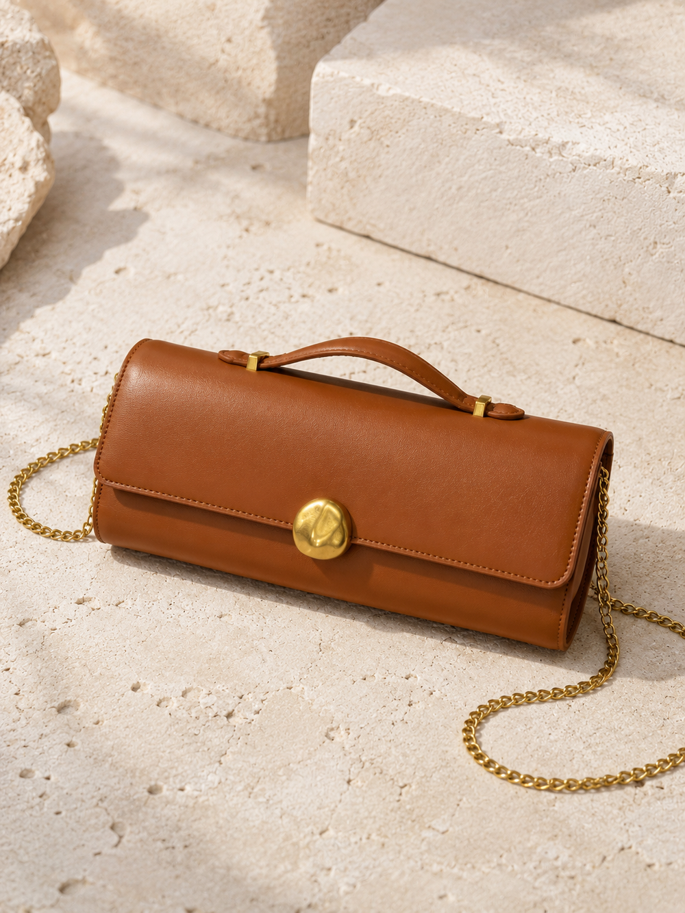 Brown leather handbag with gold chain on a light stone surface. Tan Top Handle Clutch Bag. 