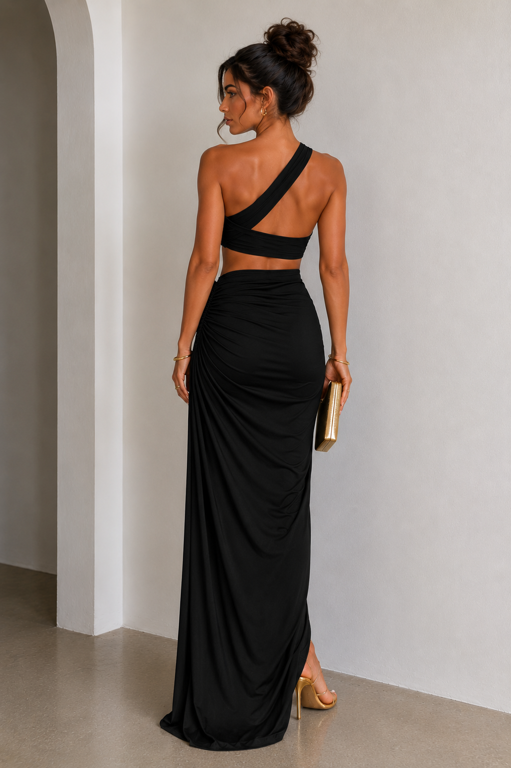 A woman modeling a black maxi dress from the back in a minimalist indoor setting. The dress features a single crossover shoulder strap and a high-waisted, draped skirt with a side slit.