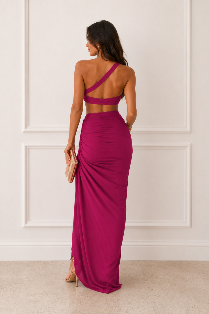A woman stands with her back to the camera wearing a magenta one-shoulder maxi dress with a cutout back and draped skirt, holding a small beige clutch against a white paneled wall.