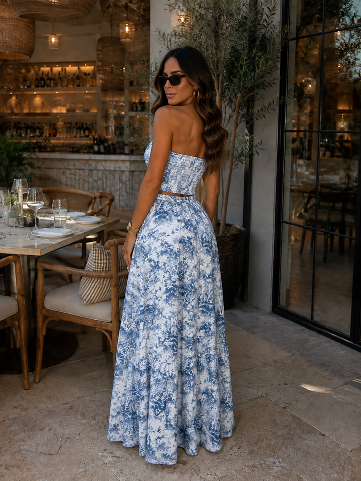 A woman stands on a restaurant patio wearing a strapless blue and white floral maxi dress, photographed from the back. The setting includes stone flooring, dining tables, chairs, and a softly lit interior background.