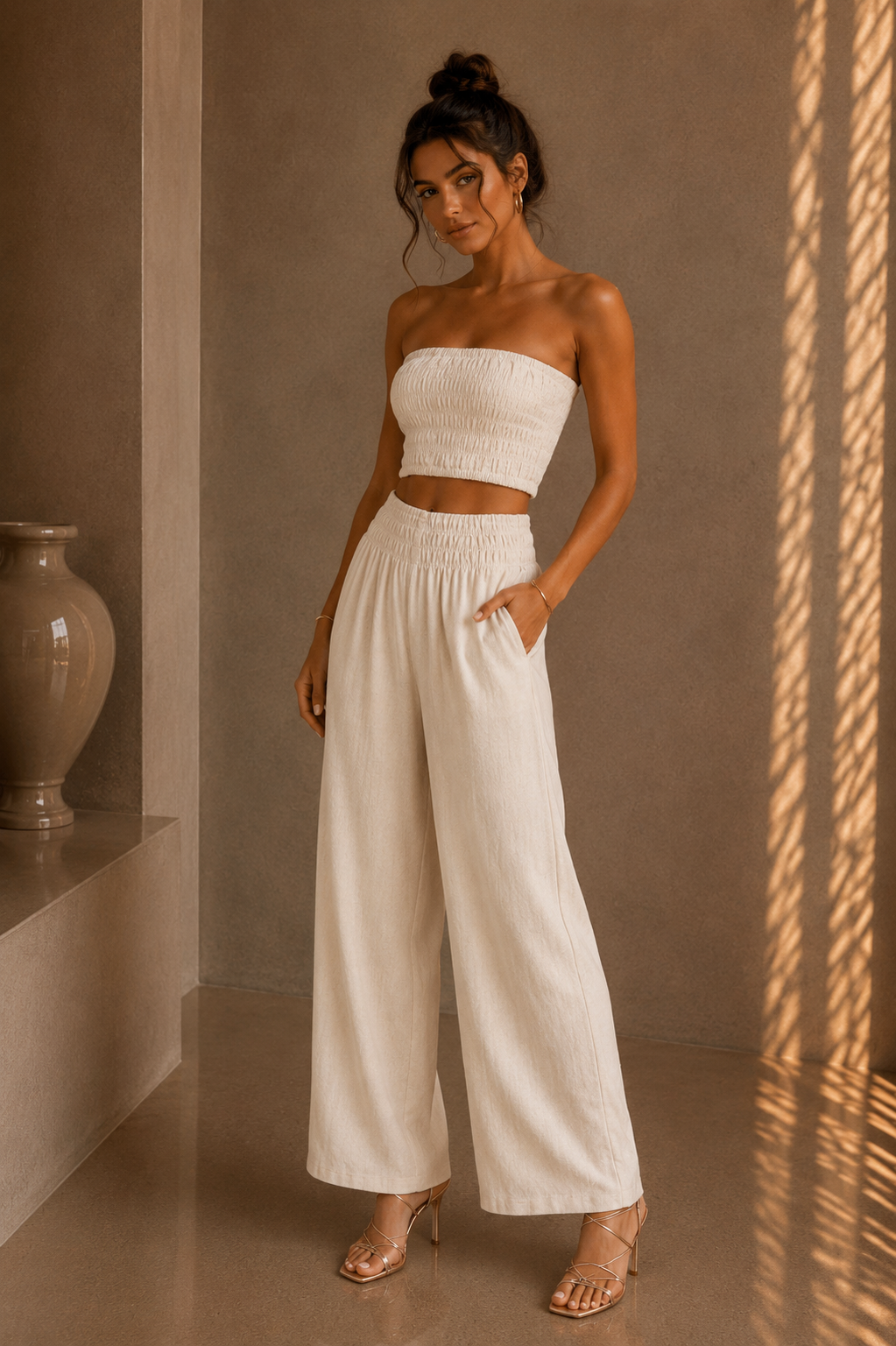Woman wearing a white strapless top and wide-leg pants standing against a beige wall.