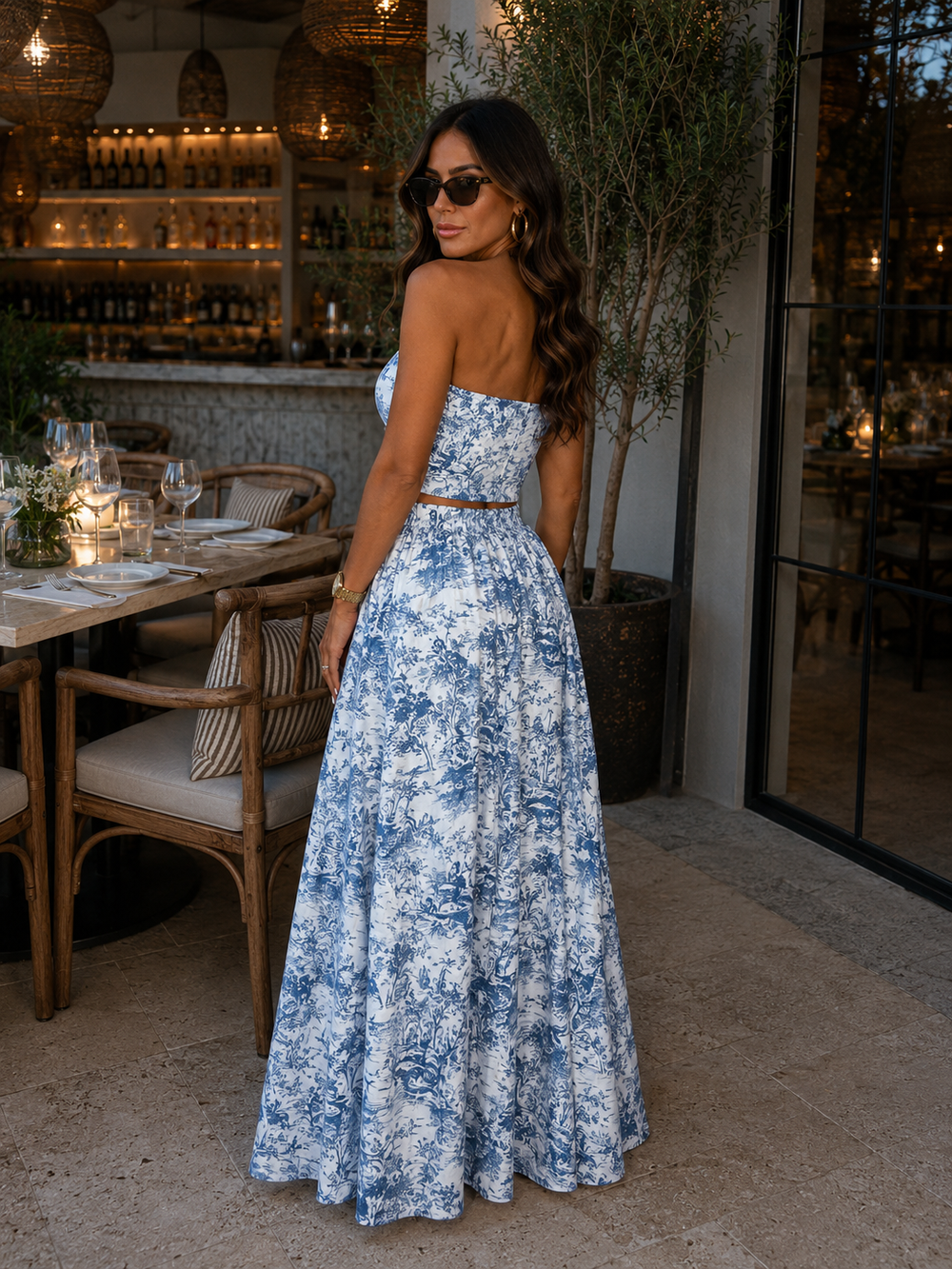 A woman wearing a strapless blue and white floral maxi dress with a matching waist belt, standing in a restaurant patio setting.