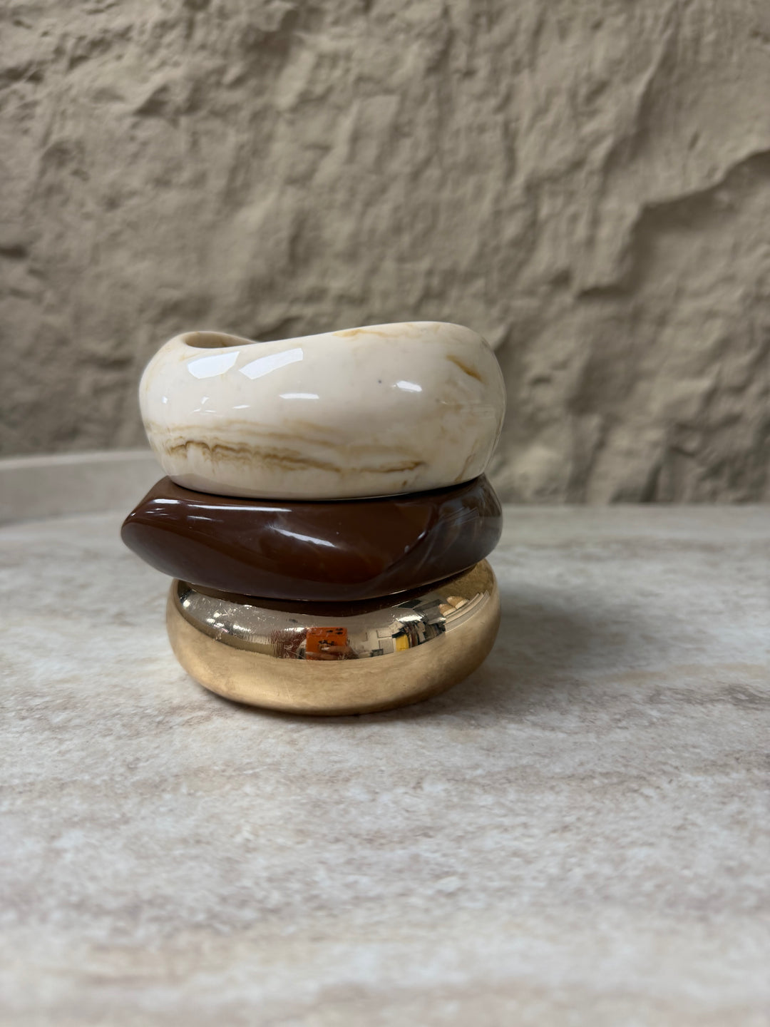 Three stacked ceramic pots in white, brown, and gold on a textured surface with a stone wall background. 