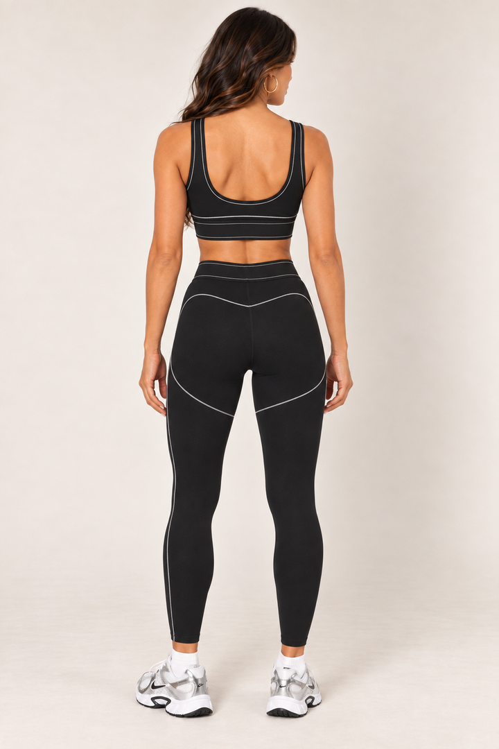 Back view of the black sports bra showing the deep scoop back, wide straps and light contrast stitching, styled with matching black leggings.
