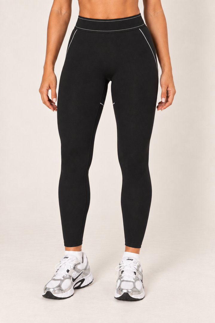 Waist-down front view of black fitted leggings showing the wide high-rise waistband, light contrast seams at the hips and smooth ankle-length legs.