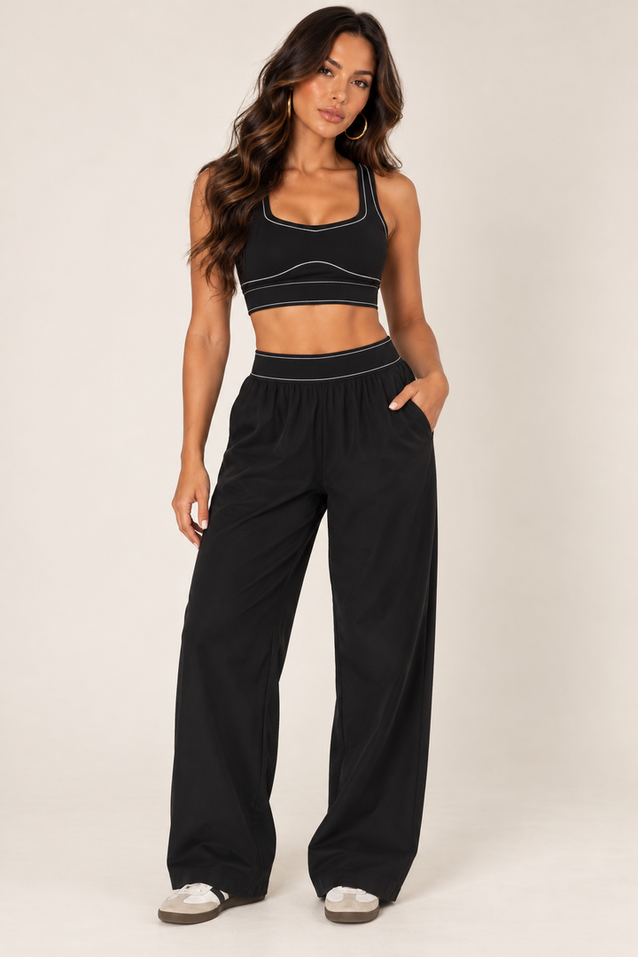 Front outfit view of Glowie black contrast-stitch sports bra paired with coordinating wide-leg black pants and sneakers, sold separately.