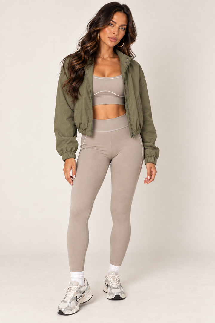 Olive LOVE TREE cropped windbreaker worn open over a taupe sports bra and contour leggings, with gold hoops and white and silver sneakers.