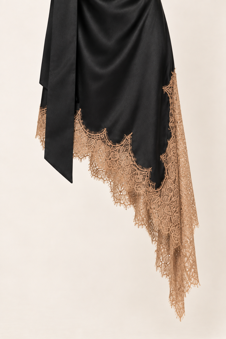 Product-only close-up of the Black Satin Scarf Midi Dress hem at Styled by ALX. Warm tan floral eyelash lace follows an irregular asymmetric black satin edge, with a long scarf tail alongside it. The ivory background shows the lace&