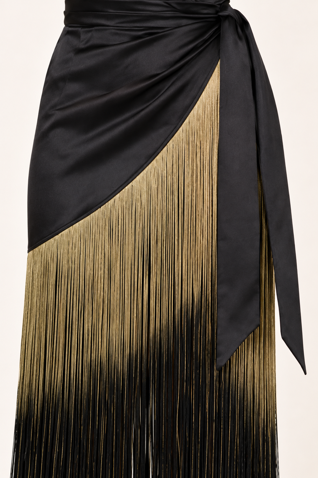 Product-only close-up of the Black Ombre Fringe Halter Dress by Skylar Rose at Styled by ALX. Fine strands of antique-gold fringe fade into black beneath the diagonal black satin skirt edge, with the matching satin waist tie alongside. The image shows the dense thread texture and attachment seam on an ivory background.