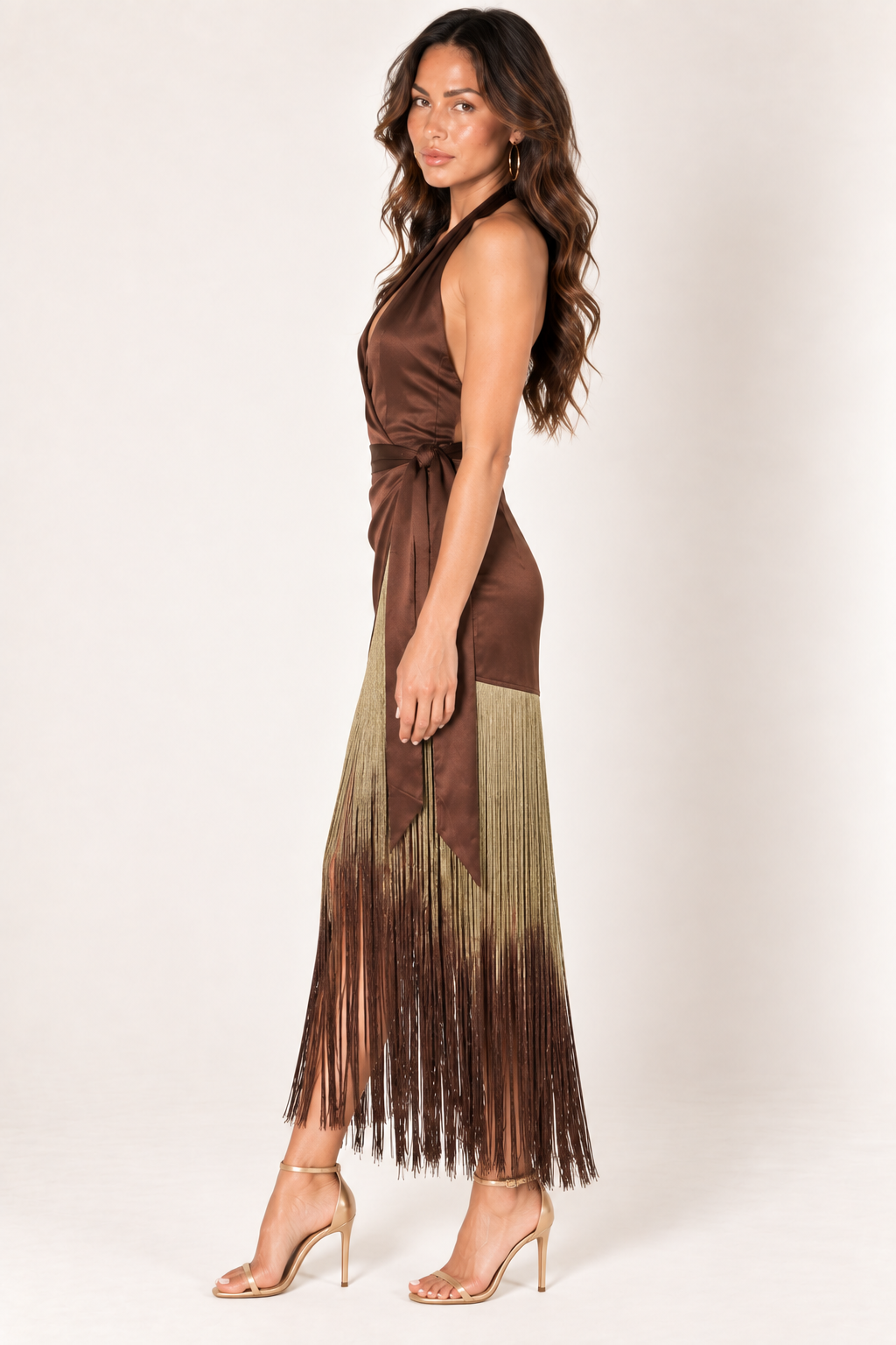 Side profile of the Brown Ombre Fringe Halter Dress by Skylar Rose at Styled by ALX. The model shows the drape of the chocolate-brown satin halter bodice, wrapped skirt panel and dense ombre fringe falling toward the ankles. Fine gold strands fade to brown at the tips above gold strappy open-toe heels.