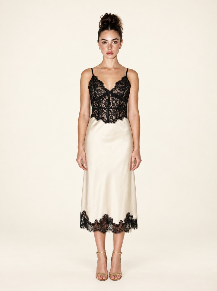 Full-length front view of the Ivory Black Lace Midi Dress by Skylar Rose at Styled by ALX. The model wears a fitted black floral lace bodice with thin straps and a scalloped neckline, an ivory satin midi skirt with black lace hem, and gold strappy open-toe heels against an ivory studio backdrop.