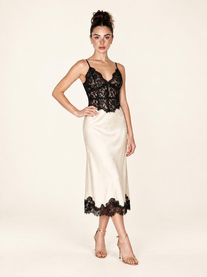 Front three-quarter view of the Ivory Black Lace Midi Dress by Skylar Rose at Styled by ALX. The angled pose shows the fitted black lace bodice, delicate shoulder straps and softly flared ivory satin skirt, with scalloped black lace at midcalf and gold strappy open-toe sandals.