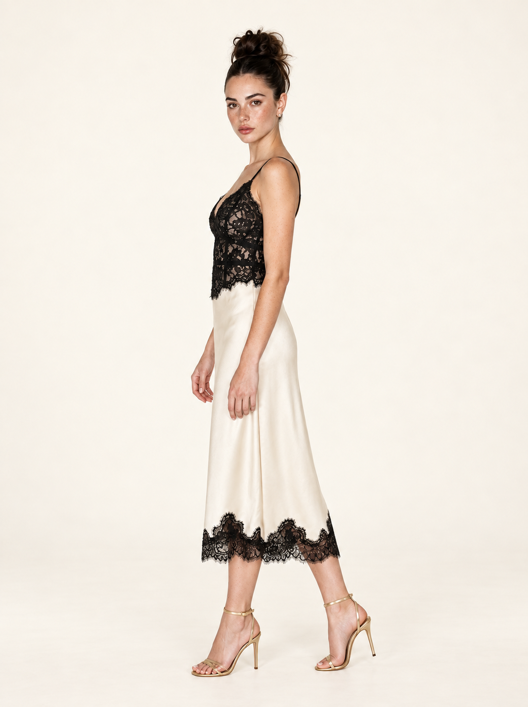 Side view of the Ivory Black Lace Midi Dress by Skylar Rose at Styled by ALX. The full-length model pose reveals the close-fitting floral lace bodice and gentle drape of the ivory satin midi skirt, ending in scalloped black lace above gold strappy open-toe heels in a warm ivory studio.