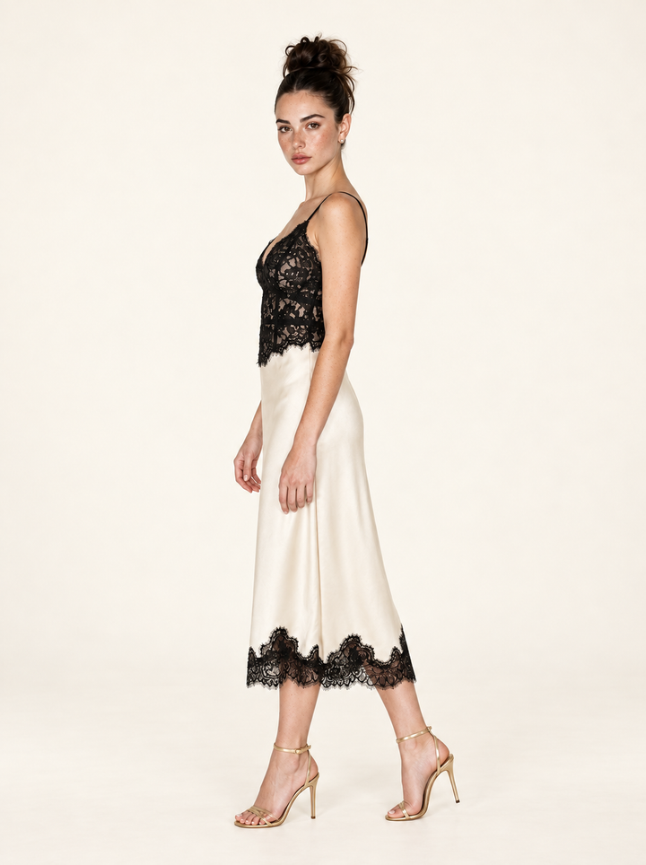 Side view of the Ivory Black Lace Midi Dress by Skylar Rose at Styled by ALX. The full-length model pose reveals the close-fitting floral lace bodice and gentle drape of the ivory satin midi skirt, ending in scalloped black lace above gold strappy open-toe heels in a warm ivory studio.