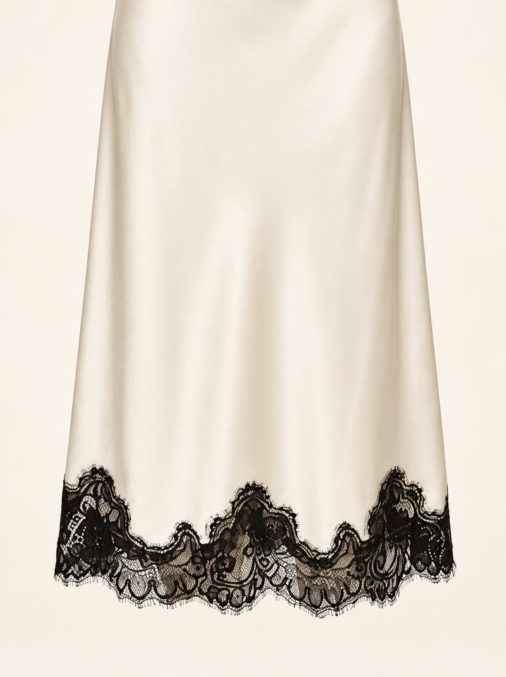 Product-only hem detail of the Ivory Black Lace Midi Dress by Skylar Rose at Styled by ALX. A close view of the ivory satin skirt shows its smooth sheen and the wide, irregular scalloped border of black floral lace, with delicate eyelash edges visible against a pale background.