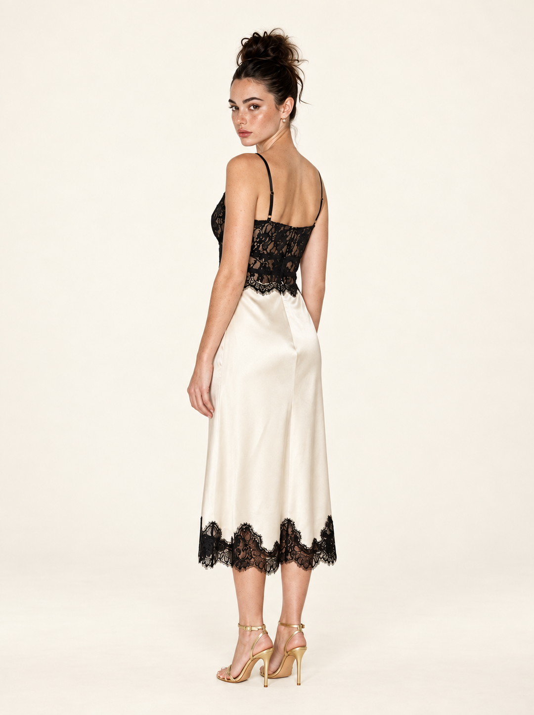 Rear three-quarter view of the Ivory Black Lace Midi Dress by Skylar Rose at Styled by ALX. This angle shows the thin adjustable straps, fitted black lace back and waist, and ivory satin skirt with a scalloped black lace midi hem, paired with gold strappy heels in a neutral studio.