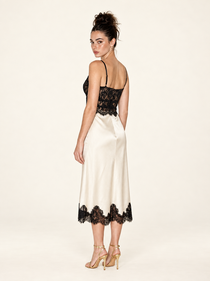 Rear three-quarter view of the Ivory Black Lace Midi Dress by Skylar Rose at Styled by ALX. This angle shows the thin adjustable straps, fitted black lace back and waist, and ivory satin skirt with a scalloped black lace midi hem, paired with gold strappy heels in a neutral studio.