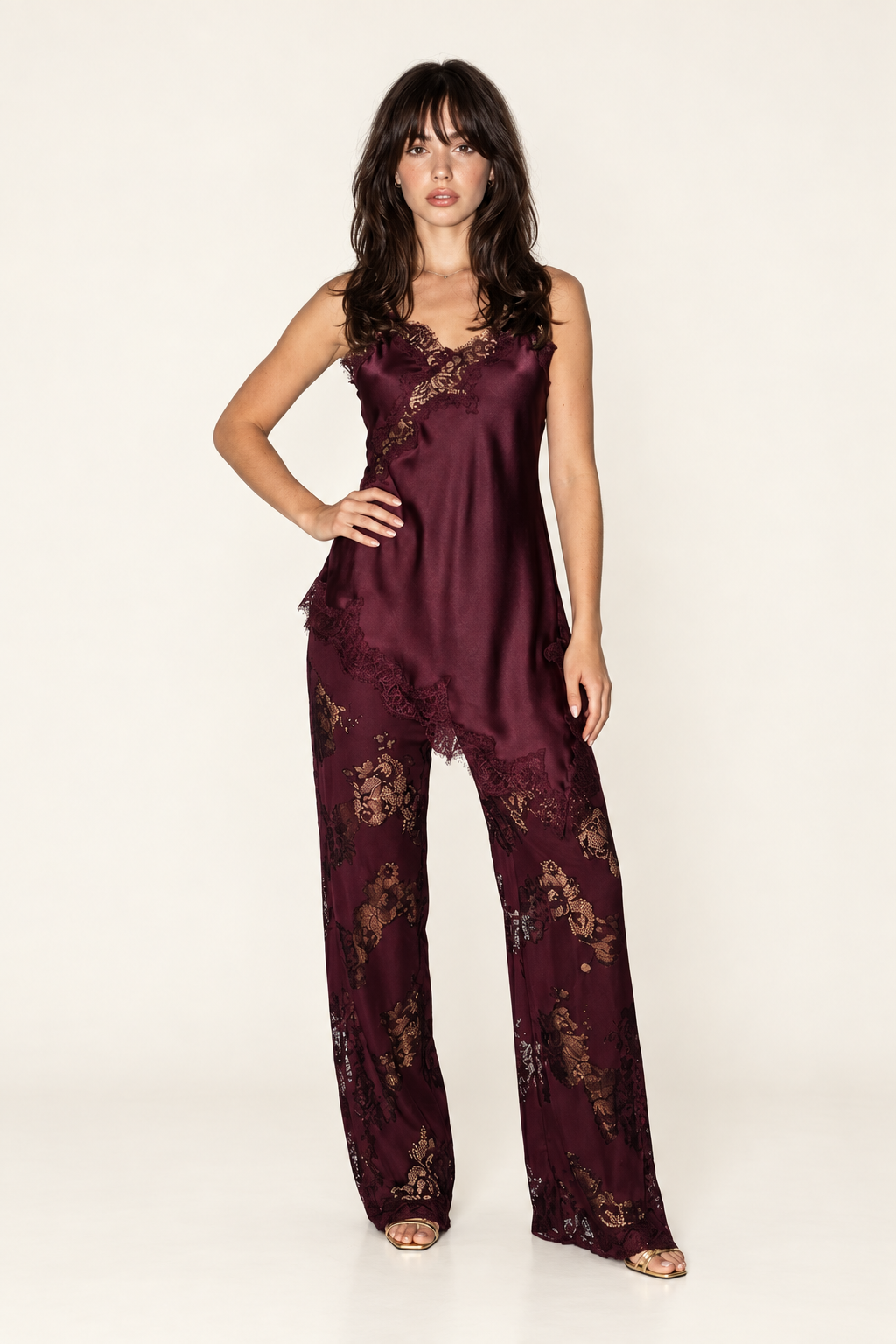 Front view of the Burgundy Satin Lace Pants Set at Styled by ALX, showing a deep wine satin camisole with spaghetti straps, a diagonal floral lace inset and an asymmetric lace-trim hem. Matching wide-leg lace pants have partial lining, styled with gold strappy open-toe heels in an ivory studio.