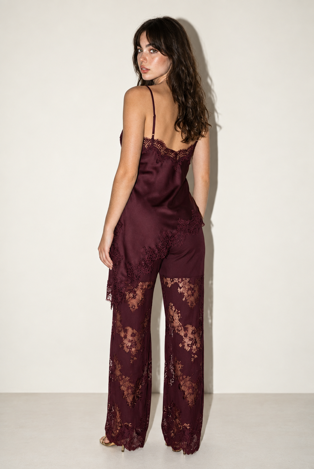 Back view of the Burgundy Satin Lace Pants Set at Styled by ALX, showing adjustable spaghetti straps, a low lace-edged camisole neckline and the asymmetric satin tunic hem. Matching wide-leg floral lace trousers have opaque upper lining, with gold strappy heels beneath the hems.