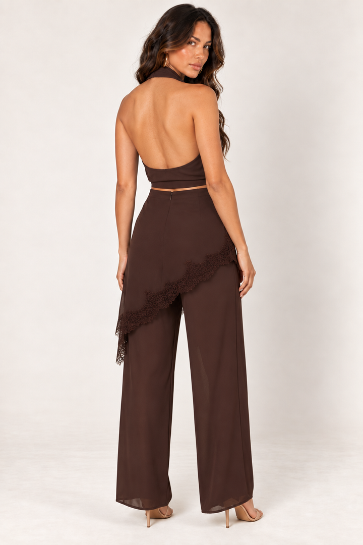 Back view of the Brown Halter Lace Pants Set at Styled by ALX. The chocolate-brown halter top reveals an open back above a smooth lower band, while the matching wide-leg pants feature a centre-back fastening and asymmetric lace-trim overlay. Styled with nude strappy heels against an ivory background.