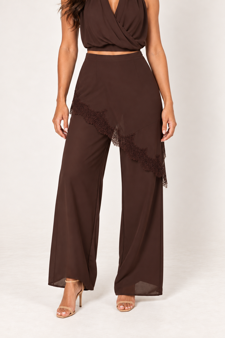 Close-up of the Brown Halter Lace Pants Set trousers at Styled by ALX, showing the smooth chocolate-brown waist, asymmetric hip overlay and delicate tonal floral lace edge above flowing wide legs. Soft ivory studio lighting highlights the layered construction, fabric drape and matching nude strappy sandals.