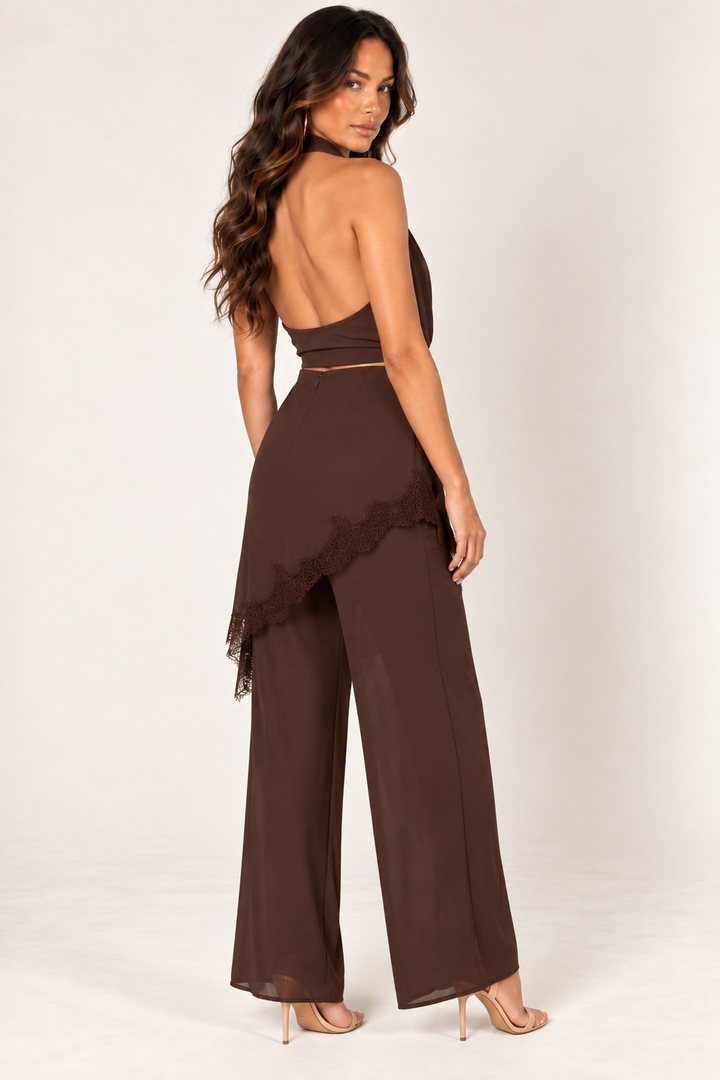 Rear three-quarter view of the Brown Halter Lace Pants Set at Styled by ALX, showing the open halter back and smooth cropped band above matching wide-leg trousers. The asymmetric chocolate-brown overlay ends in tonal lace trim, and nude strappy sandals complete the full-length studio look.